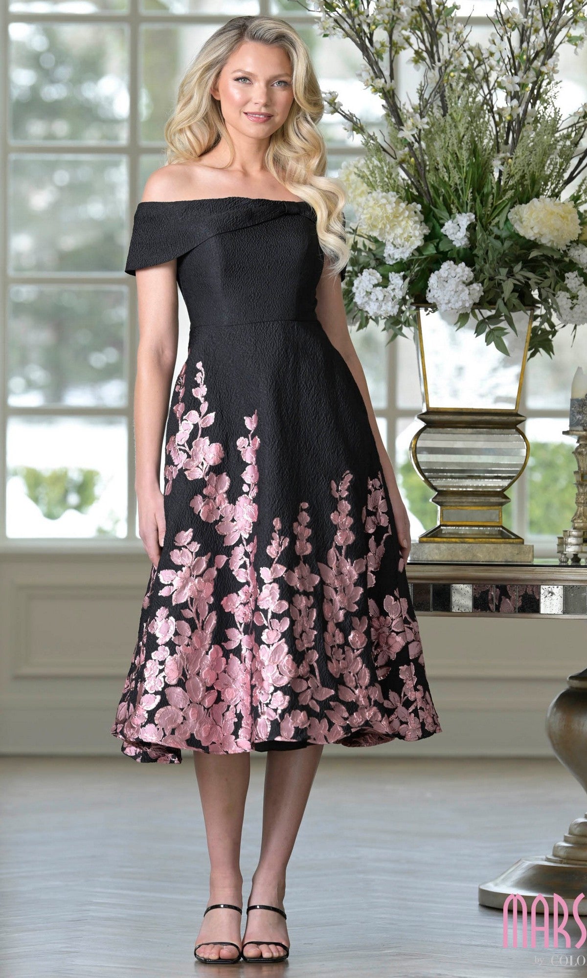 A woman stands indoors in a Marsoni MV1414 knee length semi-formal dress in rose black, paired with black heels and styled with long wavy hair, next to a large floral arrangement.